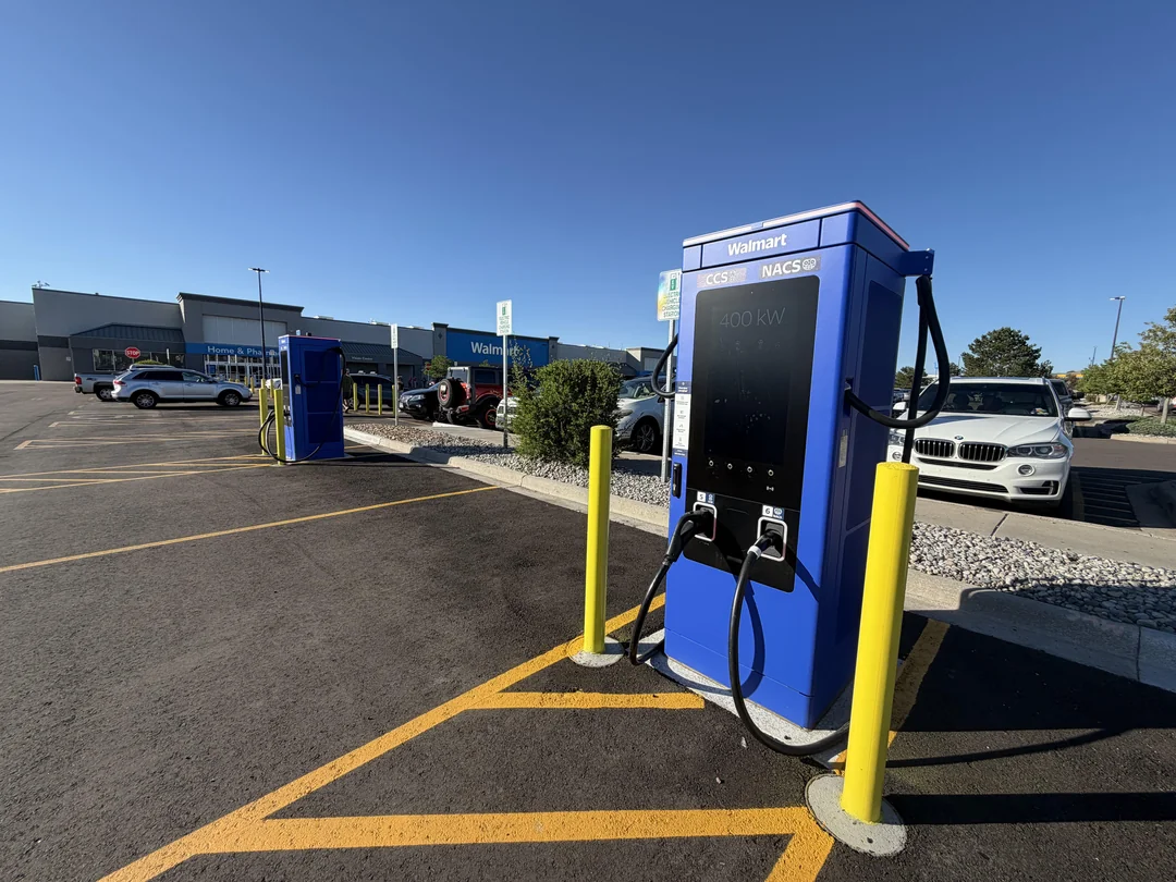 Two cobalt-blue Walmart-branded 400 kW EV fast chargers with CCS and NACS connectors stand in a Walmart parking lot under a clear blue sky, with yellow safety bollards and a white BMW SUV parked nearby.