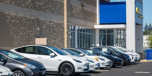 A row of used Tesla and other electric vehicles parked in the lot of a CarMax dealership on a sunny day, with white sales stickers in the windows.