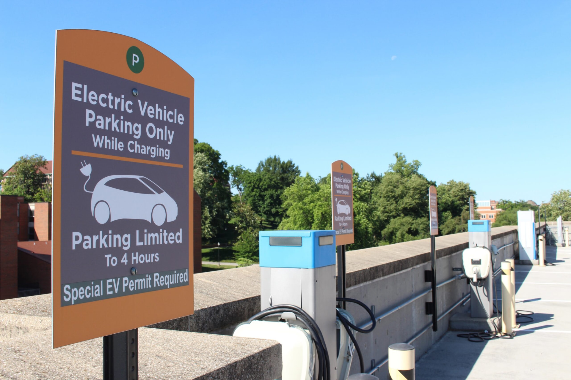 A row of EV charging stations mounted along a concrete barrier in a parking garage, with orange EV-only parking signs designating the spots under a clear blue sky.